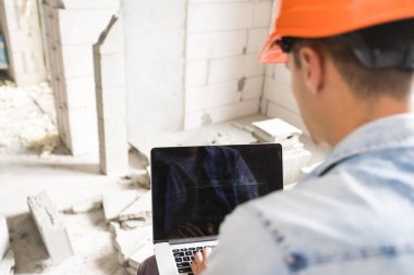 Kasklı yakışıklı bir mimar ya da ustabaşı binanın iç kısmındaki çizimler üzerinde dizüstü bilgisayarla çalışıyor.