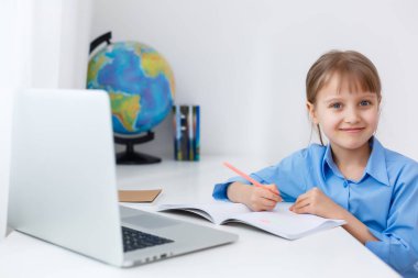 Akıllı küçük kız oturma odasında ödevini yapıyor. Masasında Otururken Ders kitabında Kalem Yazar ve Dizüstü Bilgisayar Kullanır.