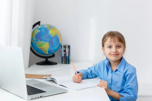Έξυπνο κοριτσάκι κάνει τις εργασίες στο σαλόνι της. Shes κάθεται στο γραφείο της γράφει με ένα στυλό στα βιβλία της και χρήσεις Laptop. — Φωτογραφία Αρχείου