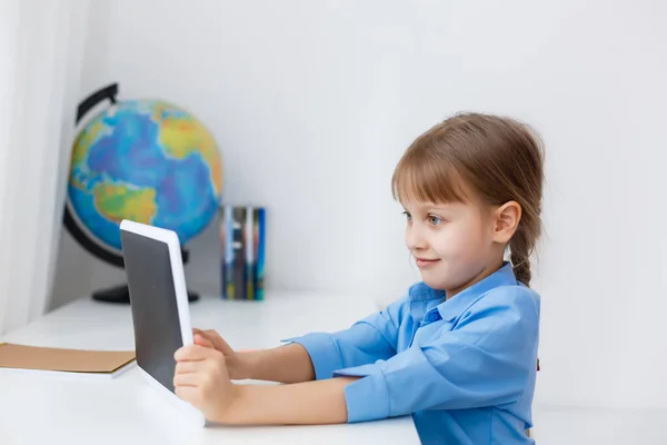 Χαριτωμένο κοριτσάκι κάθεται στο τραπέζι με το φορητό υπολογιστή της και τη μελέτη σε απευθείας σύνδεση — Φωτογραφία Αρχείου