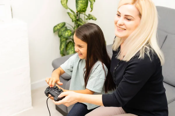 Buon rapporto carino bambina con la giovane madre utilizzando joystick giocare video gioco seduti insieme in soggiorno godendo vacanza in famiglia. — Foto Stock