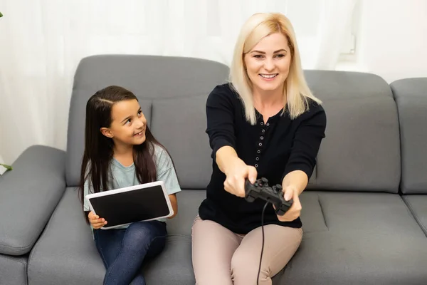 Buon rapporto carino bambina con la giovane madre utilizzando joystick giocare video gioco seduti insieme in soggiorno godendo vacanza in famiglia. — Foto Stock