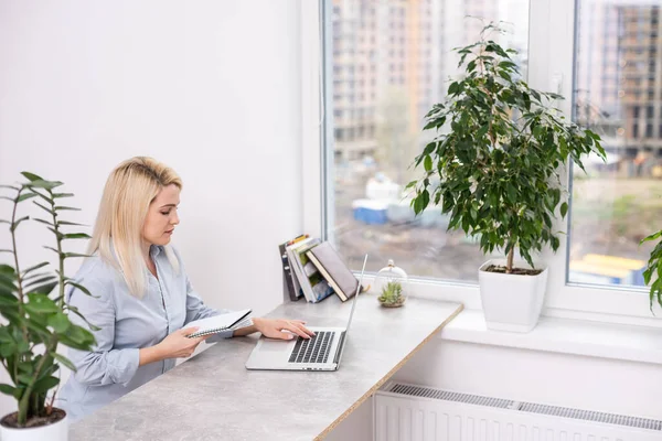 Jovem mulher branca loira fotógrafa sentada na mesa de madeira do laptop grande janela e parede cinza trabalhando — Fotografia de Stock