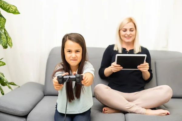 Buon rapporto carino bambina con la giovane madre utilizzando joystick giocare video gioco seduti insieme in soggiorno godendo vacanza in famiglia. — Foto Stock