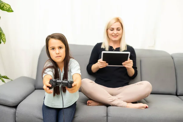 Buon rapporto carino bambina con la giovane madre utilizzando joystick giocare video gioco seduti insieme in soggiorno godendo vacanza in famiglia. — Foto Stock