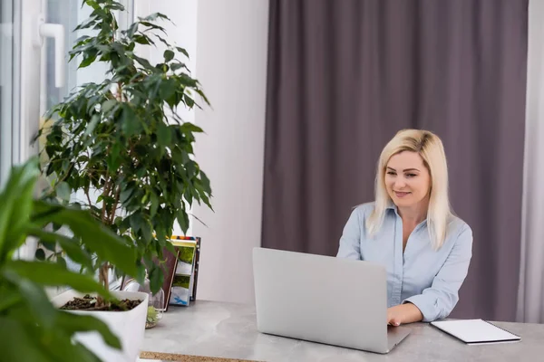 Zelfverzekerde serieuze ervaren gekwalificeerde mooie slimme vrouw met blond haar in shirt typt brieven naar haar klanten en zakenpartners, zittend aan de tafel in het kantoor — Stockfoto