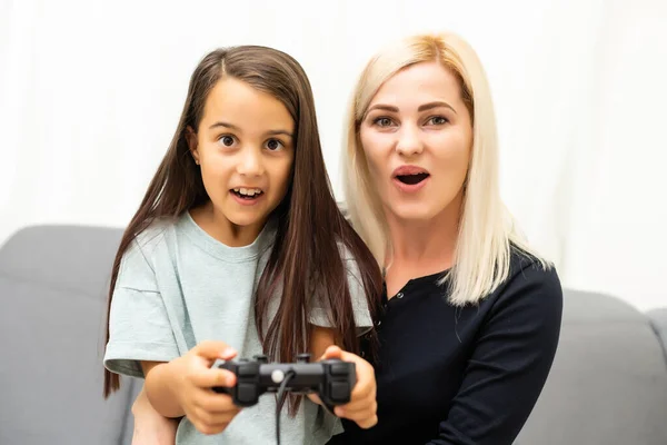 Buon rapporto carino bambina con la giovane madre utilizzando joystick giocare video gioco seduti insieme in soggiorno godendo vacanza in famiglia. — Foto Stock