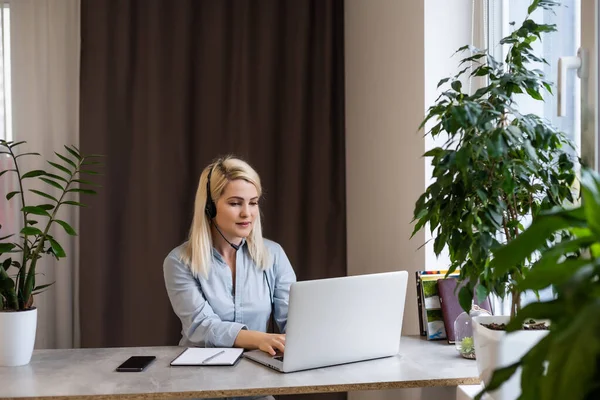 Jovem mulher branca loira fotógrafa sentada na mesa de madeira do laptop grande janela e parede cinza trabalhando — Fotografia de Stock