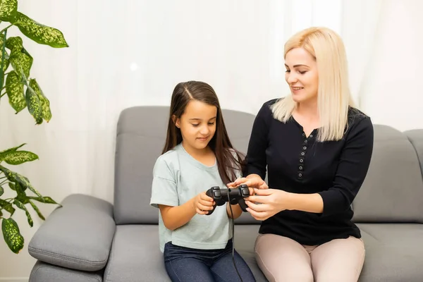 Buon rapporto carino bambina con la giovane madre utilizzando joystick giocare video gioco seduti insieme in soggiorno godendo vacanza in famiglia. — Foto Stock