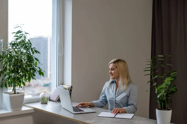 Νεαρή γυναίκα που κάθεται στο γραφείο της μπροστά από ένα γκρι φορητό υπολογιστή ενώ ονειροπολεί όμορφες αναμνήσεις ή το μέλλον σε ένα σύγχρονο φωτεινό γραφείο με μεγάλο παράθυρο — Φωτογραφία Αρχείου
