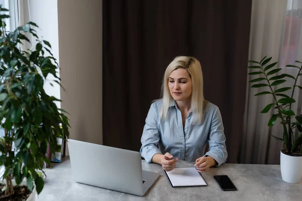 Jonge vrouw zit achter haar bureau voor een grijze laptop terwijl ze dagdroomt van mooie herinneringen of toekomst in een modern helder kantoor met groot raam — Stockfoto