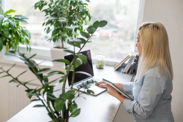 Jonge vrouw zit achter haar bureau voor een grijze laptop terwijl ze dagdroomt van mooie herinneringen of toekomst in een modern helder kantoor met groot raam — Stockfoto