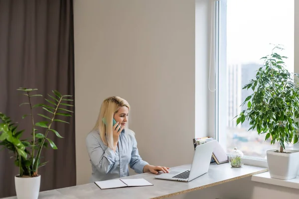 Jovem mulher sentada em sua mesa na frente de um laptop cinza enquanto sonha acordado com boas memórias ou futuro em um escritório moderno brilhante com grande janela — Fotografia de Stock