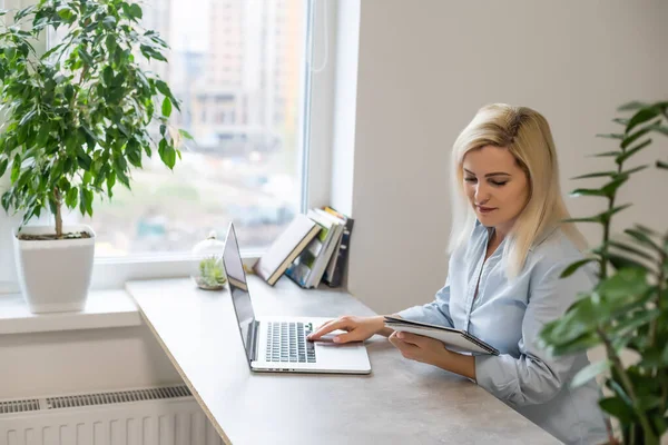 Уверенная в себе серьезная опытная квалифицированная красивая умная женщина с светлыми волосами в рубашке печатает письма своим клиентам и деловым партнерам, сидя за столом в офисе — стоковое фото