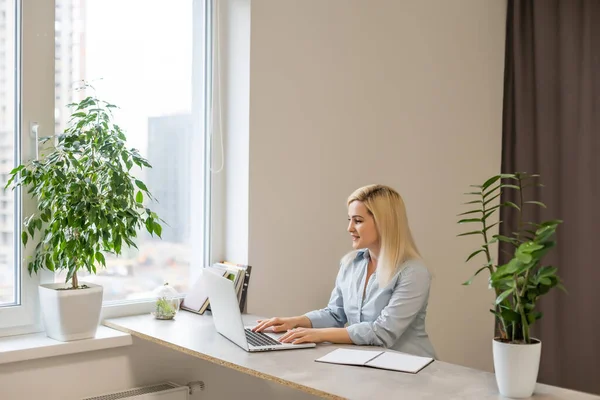 Junge Frau sitzt an ihrem Schreibtisch vor einem grauen Laptop und träumt von schönen Erinnerungen oder Zukunft in einem modernen, hellen Büro mit großem Fenster — Stockfoto