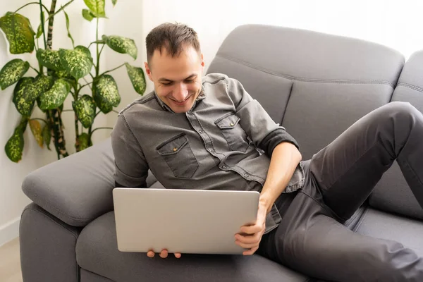 Joyeux jeune homme en chemise sur canapé à la maison, travaillant sur ordinateur portable, souriant. — Photo