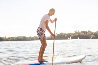 Plajda SUP sörf tahtası olan bir sörfçünün portresi. Şafakta kürek çeken genç bir adam. Ekstrem spor kavramı. Erkek sörfçü yaşam tarzı.