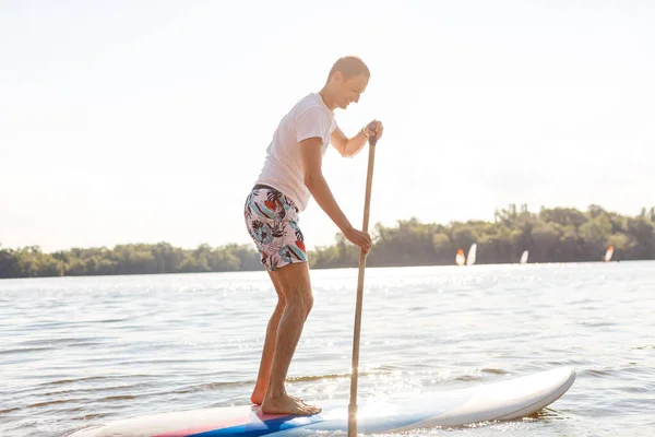 ビーチにSUPボードを持つサーファーの肖像画。夜明けにパドルボードの若い男。極端なスポーツの概念。男性サーファーのライフスタイル. — ストック写真