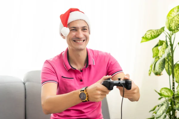 Junger Mann mit roter Neujahrsmütze, der im Wohnzimmer Videospiele mit Joystick spielt. Weihnachtsspaß, allein zu Hause genießen — Stockfoto