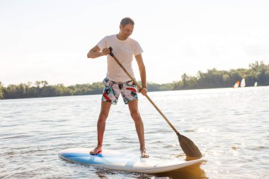 Plajda SUP sörf tahtası olan bir sörfçünün portresi. Şafakta kürek çeken genç bir adam. Ekstrem spor kavramı. Erkek sörfçü yaşam tarzı.