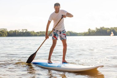 Plajda SUP sörf tahtası olan bir sörfçünün portresi. Şafakta kürek çeken genç bir adam. Ekstrem spor kavramı. Erkek sörfçü yaşam tarzı.
