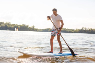 Plajda SUP sörf tahtası olan bir sörfçünün portresi. Şafakta kürek çeken genç bir adam. Ekstrem spor kavramı. Erkek sörfçü yaşam tarzı.