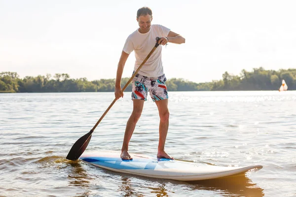 Portret surfera z tablicą SUP na plaży. Młody człowiek na desce o świcie. Koncepcja sportów ekstremalnych. Mężczyzna surfer styl życia. — Zdjęcie stockowe