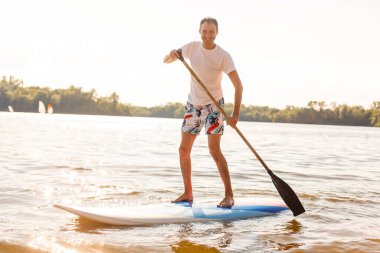 Plajda SUP sörf tahtası olan bir sörfçünün portresi. Şafakta kürek çeken genç bir adam. Ekstrem spor kavramı. Erkek sörfçü yaşam tarzı.