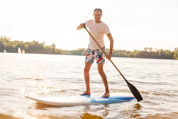 Portrait of a surfer with a SUP Board on the beach. Young man on paddleboard at dawn. The concept of extreme sports. Male surfer lifestyle. — Stock Photo, Image