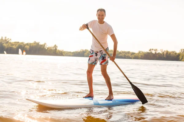 Retrato de un surfista con una tabla SUP en la playa. Joven en el paddleboard al amanecer. El concepto de deportes extremos. Estilo de vida del surfista masculino. — Foto de Stock