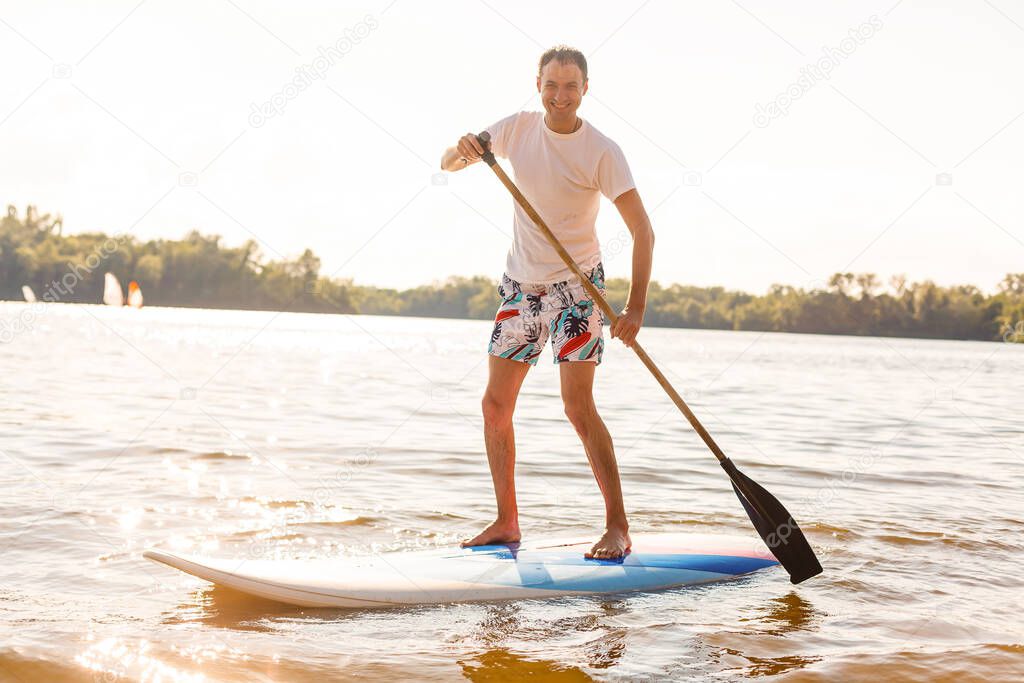 Portrait of a surfer with a SUP Board on the beach. Young man on paddleboard at dawn. The concept of extreme sports. Male surfer lifestyle.