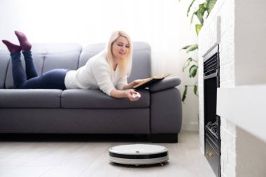 Kadın kanepede dinlenirken robot süpürge odayı temizliyor. Uzaktan kumandayla vakumu kontrol eden kadın