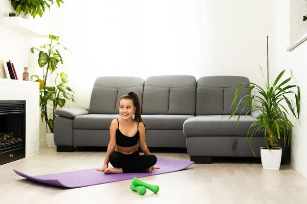 La niña está haciendo ejercicios de sentadilla en casa. Lindo niño está entrenando en una alfombra interior. Pequeña modelo femenina de pelo oscuro en ropa deportiva tiene ejercicios cerca de la ventana en su habitación — Foto de Stock