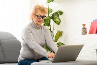 Evde dizüstü bilgisayarıyla gülümseyen yaşlı bir kadın.