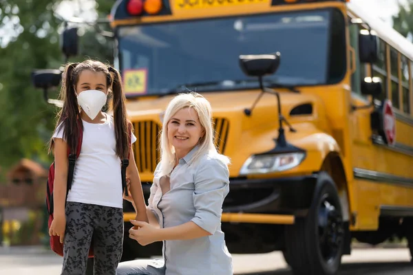 Caucasian woman puts a protective mask on her daughter outdoors. Caring mother helps to wear a mask schoolgirl near the school bus. Quarantine during coronavirus.