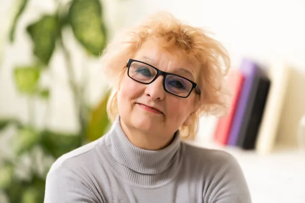 Sonriente de mediana edad madura mujer de pelo gris mirando a la cámara, feliz edad posando en casa interior, positivo solo senior mujer jubilada sentada en el sofá en la sala de estar retrato de la cabeza —  Fotos de Stock