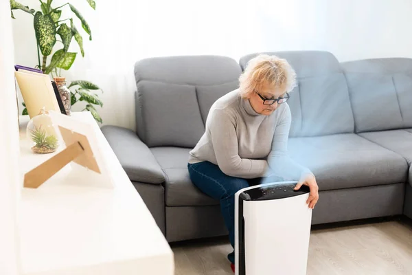 Una mujer de edad avanzada utiliza un purificador de aire en un apartamento, mientras que la situación de contaminación del aire de polvo fuera —  Fotos de Stock