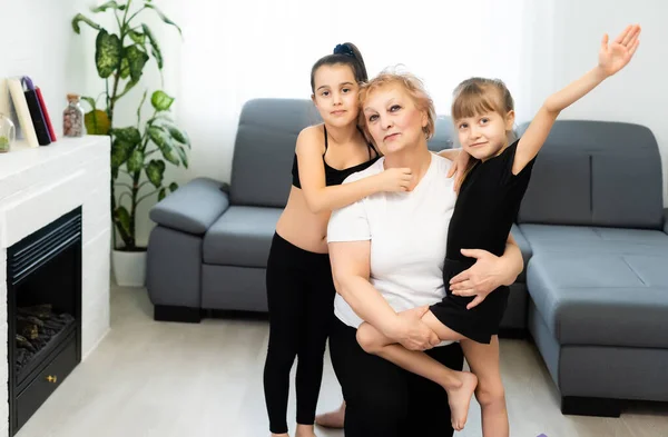 Abuela senior entrena yoga con nietas preescolares. Están sentados en la alfombra en el acogedor interior de la casa. Concepto de deporte, paternidad y personas — Foto de Stock