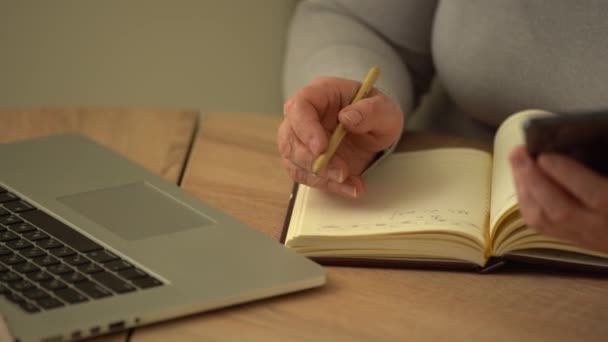 Close-up gerimpelde mannelijke handen schrijven informatie. Oude volwassen vrouw werkt op kantoor, met behulp van computer, handschrift notities — Stockvideo