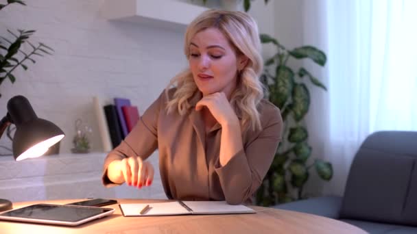 Χαμογελώντας ελκυστική γυναίκα σκέφτεται κάτι στη συνέχεια, γράφοντας κάτι στο σημειωματάριο, ενώ κάθεται στο τραπέζι — Αρχείο Βίντεο