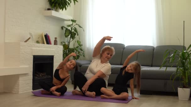 Volwassen vrouw met haar kleindochters. Ochtend oefeningen, yoga - doe thuis allemaal samen. Grootmoeder en kleindochters doen yoga oefening thuis. Moeder en dochter doen gymnastiek oefeningen. — Stockvideo