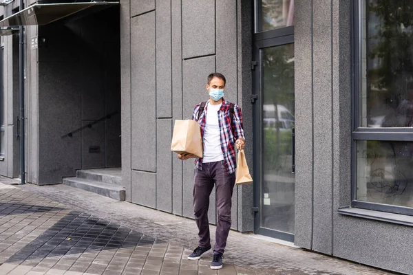 Homem de entrega jovem em máscara médica no rosto segurando sacos de papel nas mãos. Correio Hipster com sacos de compras. Serviço de entrega online seguro. Proteção contra o Coronavirus COVID-19 — Fotografia de Stock