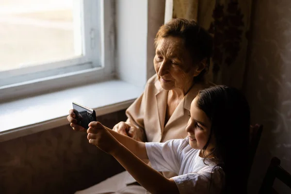 증조할머니는 증손녀와 함께 앉아서 스마트폰을 들여다 봅니다. 할머니와 아이들은 스마트폰으로 셀카를 먹습니다. 손녀딸과 함께 전화기를 들여다보는 할머니. — 스톡 사진
