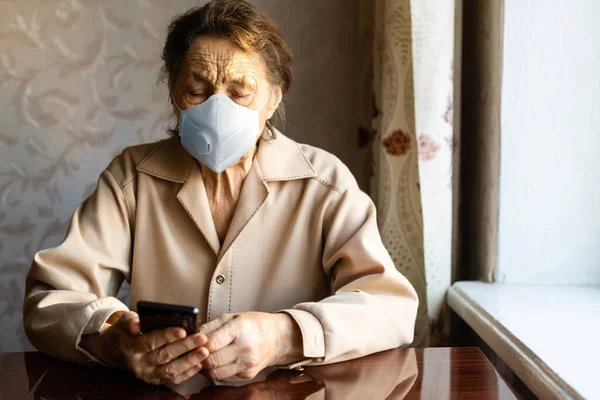 Une femme âgée dans un masque près de la fenêtre avec un smartphone — Photo