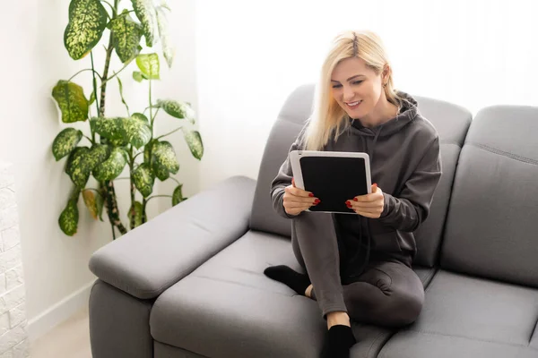 Günlük giysiler içinde güzel bir kadın dijital tablet kullanıyor ve evde gülümsüyor. — Stok fotoğraf