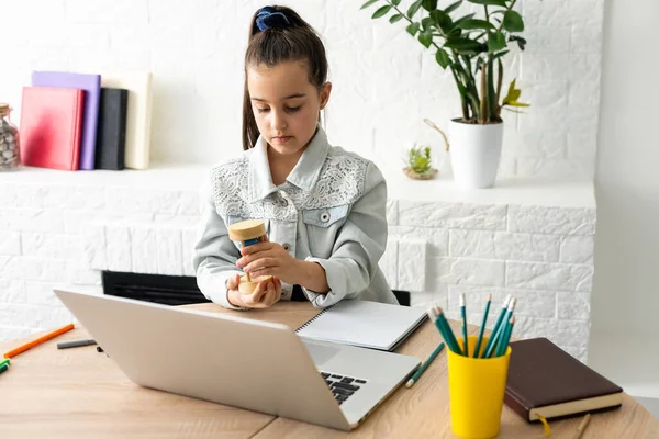 Klein meisje zit met een laptop aan tafel en houdt een zandloper, wachten op het einde van online lessen — Stockfoto