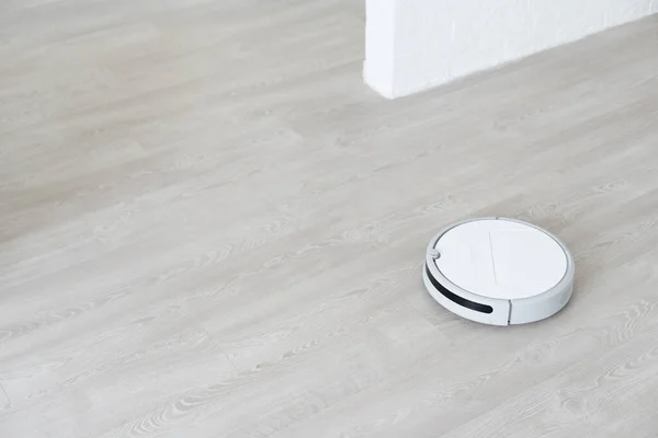 White robotic vacuum cleaner on laminate floor cleaning dust in living room interior. Smart housekeeping technology.