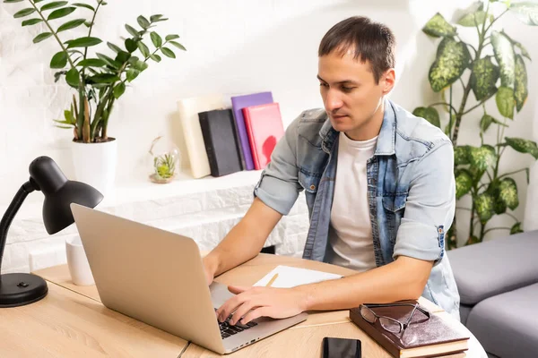 Portrait d'un homme travaillant à la maison sur un projet, il est assis à une table, dessinant des idées sur un carnet, avec son ordinateur portable devant lui. — Photo