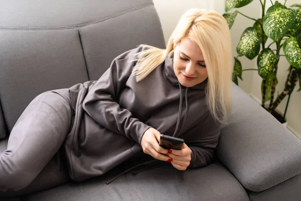 Framifrån på ung kvinna sitter hemma på soffan och använder smartphone. Flickan använder digitala prylar. Kvinnliga online shopping, surfa internet, chatta. Frilansare som arbetar hemma. — Stockfoto