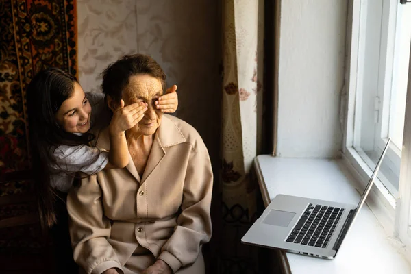 Счастливая старшая женщина сидит со своей внучкой и смотрит на ноутбук, делая видеозвонок. Зрелая леди разговаривает с веб-камерой, ведет онлайн-чат дома во время самоизоляции. Семейное время в Короне — стоковое фото